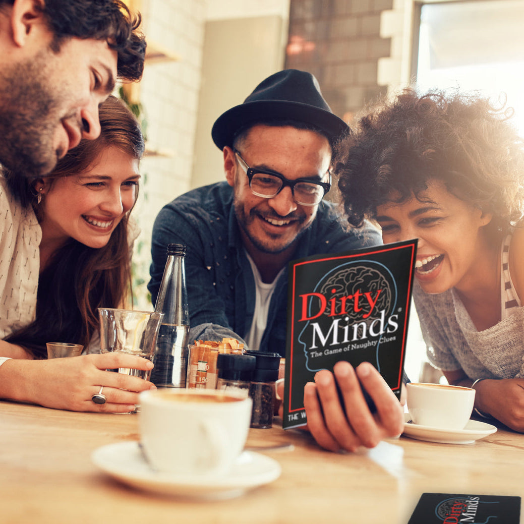 Four people gathered around a table in a warm, sunlit indoor setting, laughing while looking at a card game box titled "Dirty Minds" held by a hand in the foreground.