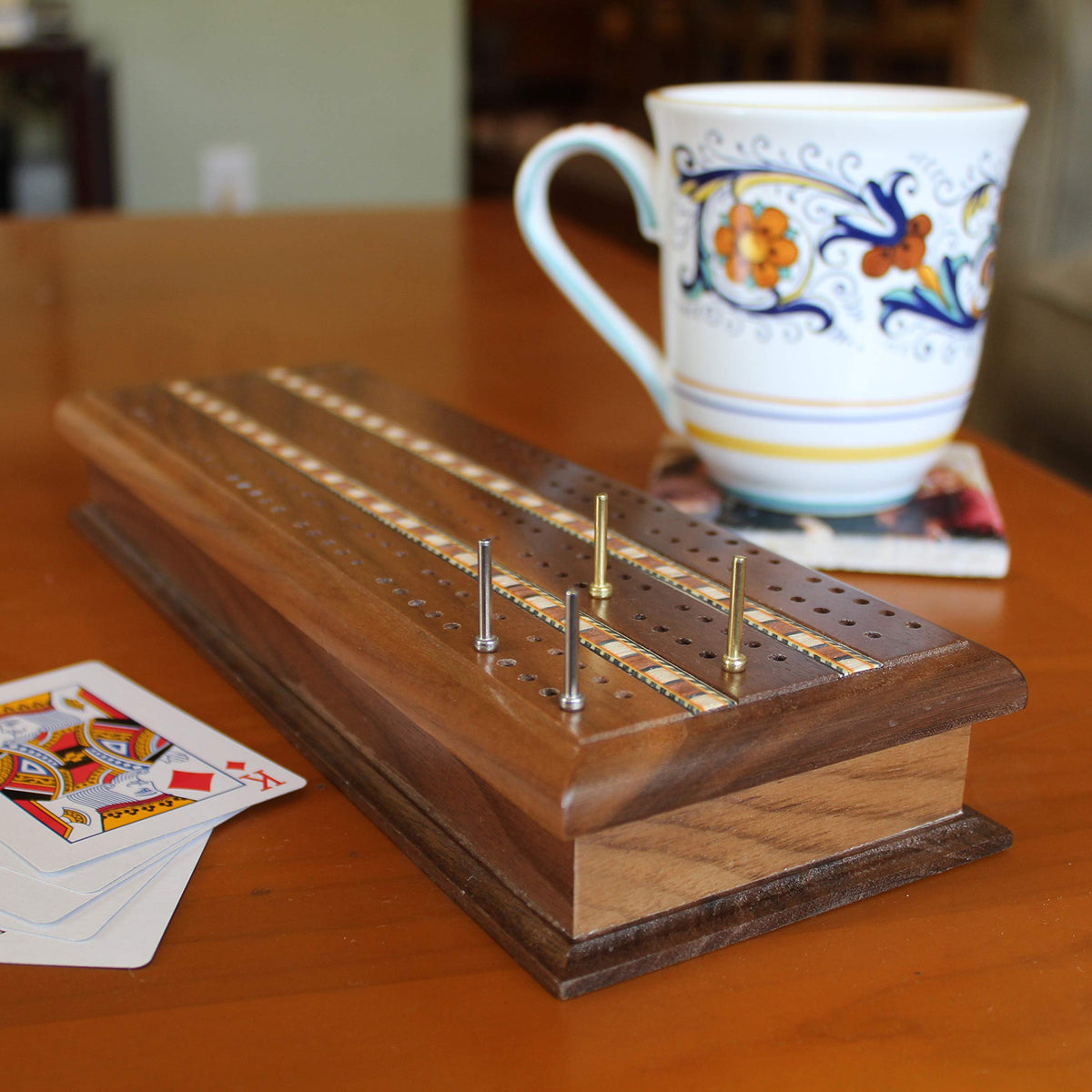 Wooden cribbage board with pegs on a table next to a decorative mug and playing cards.