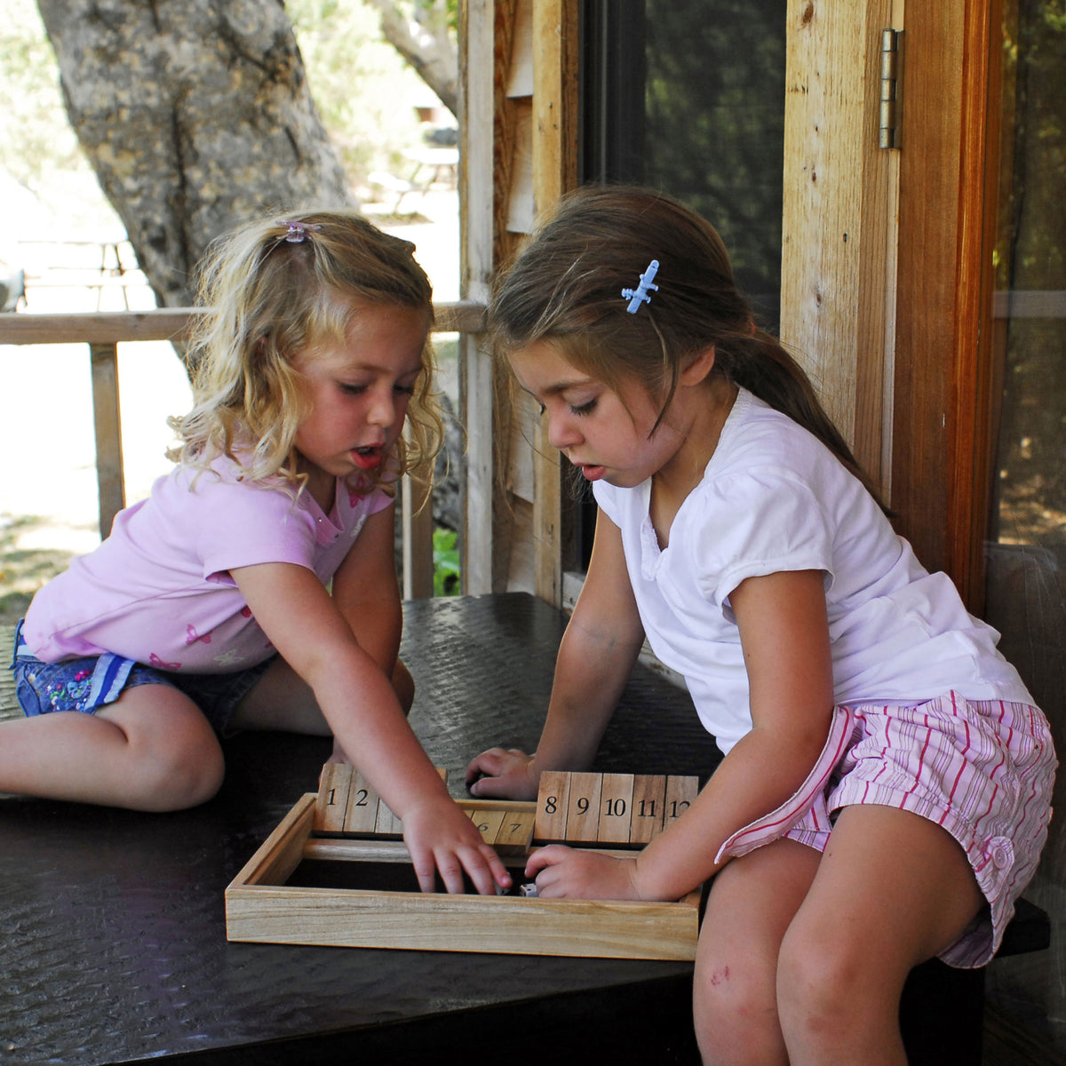 Two young girls playing with a wooden puzzle on a table outdoors.