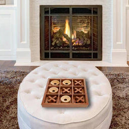 A wooden square tic-tac-toe game board with circular discs and cross markers sits on a round white tufted ottoman in front of a fireplace.