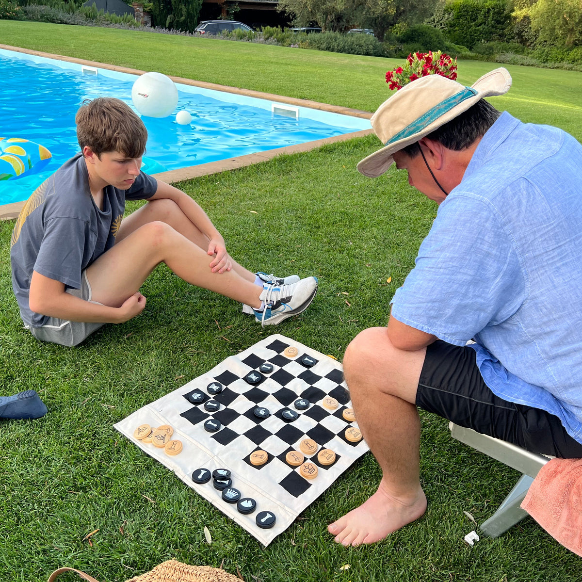 A man and a young man sitting on a grassy lawn by a swimming pool, playing a game of checkers on a white cloth. The man wears a light blue shirt, black shorts, and a straw hat, while the young man wears a gray t-shirt and white sneakers.