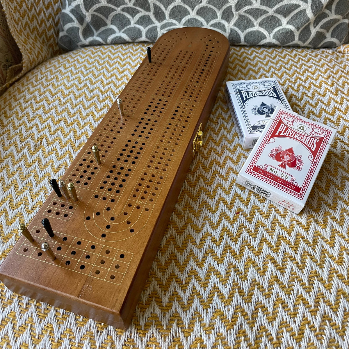 A wooden cribbage board game set on a patterned armchair, featuring a long wooden board with holes and pins, alongside two decks of playing cards.