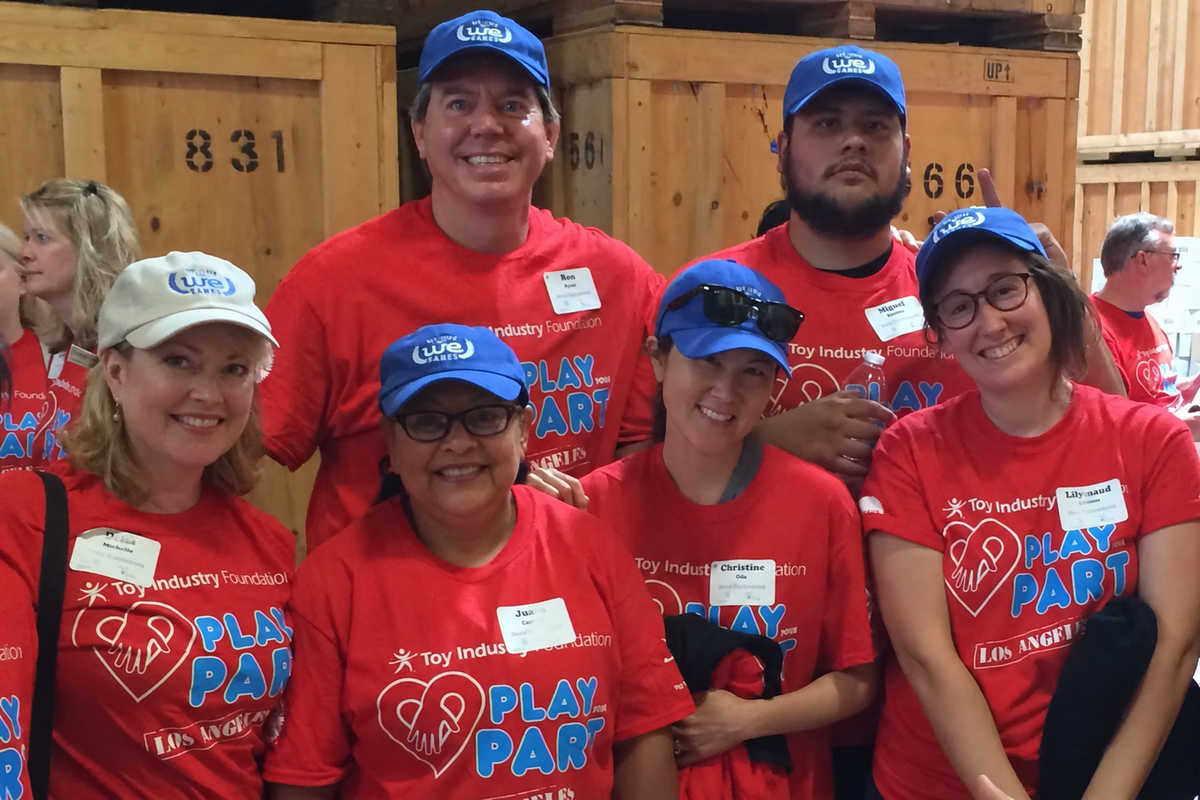 A group of seven people wearing matching red 'Toy Industry '98' t-shirts and blue 'USA' baseball caps, smiling in front of wooden storage bins. The shirts feature a white heart logo and the text 'PLAY PART'.
